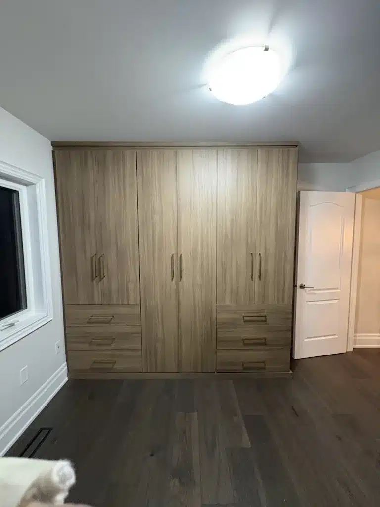 A built-in wardrobe run in a wood-look finish, built wall to wall in an Ontario bedroom with no closet, with tall door bays and a bank of drawers at each end.