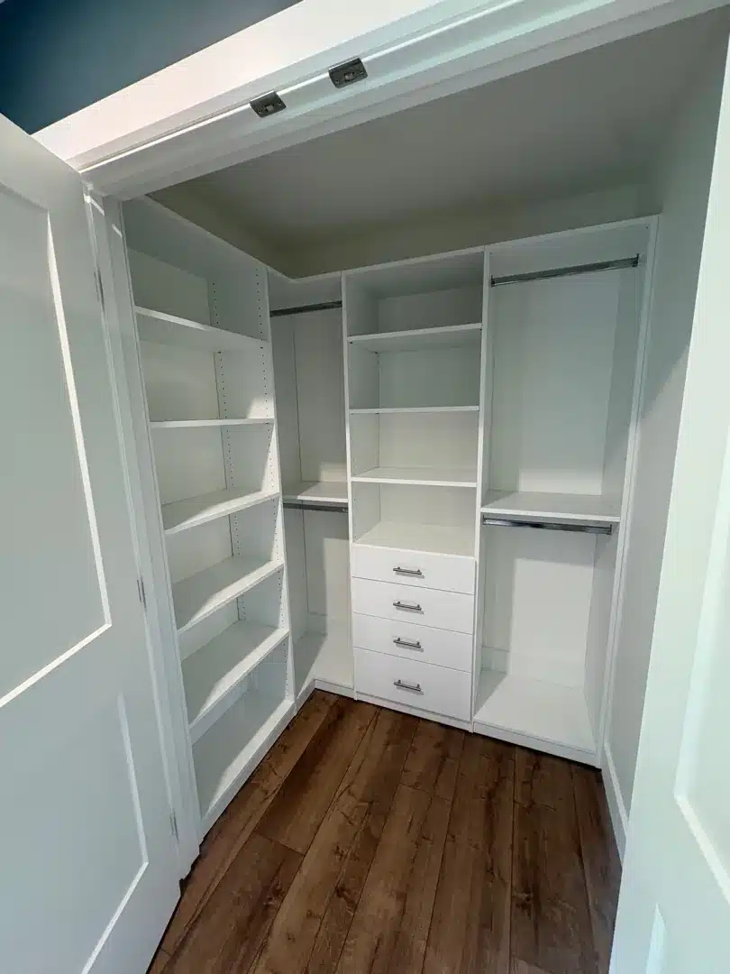 L-shaped white walk-in closet in Carling with a shelf tower, four-drawer stack and hanging rods along both walls